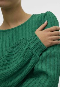 Grøn bluse med tekstur og ribbet mønster, rund halsudskæring og lange, voluminøse ærmer. Stoffet ser blødt og let krøllet ud.