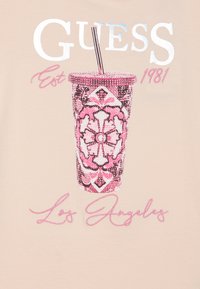 Bež majica s grafikom koja prikazuje ružičasti, ukrašeni čašu za piće ukrašenu kristalima, okruženu stiliziranim natpisom "GUESS Est 1981 Los Angeles."