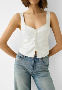 Bílý saténový top s krátkými ruk�ávy, čtvercovým výstřihem a předními knoflíky. V kombinaci se světle modrými džínami s vysokým pasem.