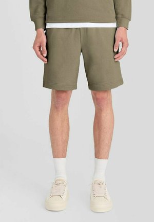 Mand iført olivengrønne shorts, hvide sokker og cremefarvede sneakers, der står mod en ensfarvet baggrund.
