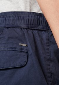 Gros plan sur un pantalon à taille élastique bleu marine avec une poche arrière et une petite étiquette métallique de la marque portant l'inscription « SOLID ».