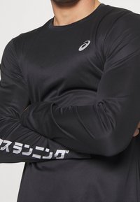Schwarzes Langarm-Sportshirt aus glattem, feuchtigkeitsableitendem Stoff mit einem grauen Logo auf der oberen Brust und weißem Text entlang des Ärmels.