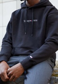 Persona vistiendo una sudadera negra de Tommy Jeans con texto del logo y pantalones grises, sentado con las manos entrelazadas sobre una superficie de piedra.