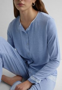 Frau sitzt und trägt ein hellblaues, langärmliges Oberteil mit Knöpfen sowie passende Hosen mit weißer Spitzenverzierung an Ärmeln und Hosenbeinen.