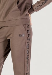 Primer plano de un chándal marrón de Emporio Armani EA7, mostrando el logo en los pantalones y en la manga, con una mano apoyada en la cadera y esmalte de uñas rojo visible.
