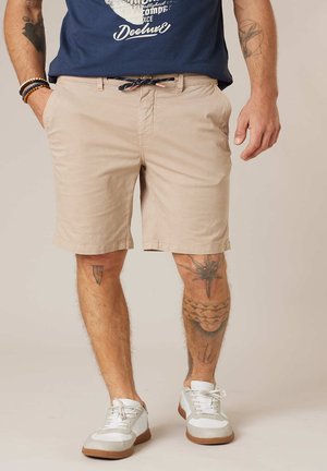 Man draagt beige korte broek met trekkoord, witte sneakers, een navy t-shirt met print, armbanden aan zijn linkerpols en tatoeages op beide benen en zijn rechterarm.