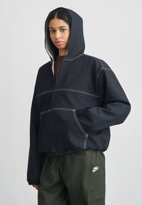 Μαύρο μπουφάν με κουκούλα και λευκές λεπτομέρειες ραφής. Εμπρόσθιο φερμουάρ, δύο πλαϊνές τσέπες, φορεμένο πάνω από χακί παντελόνι cargo με λογότυπο.