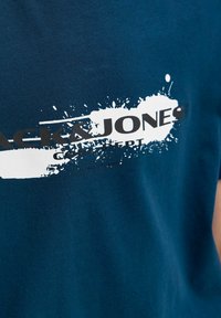 Plavi materijal s bijelim grafičkim detaljem u obliku mrlje od boje i crnim tekstom "JACK & JONES" u središtu.