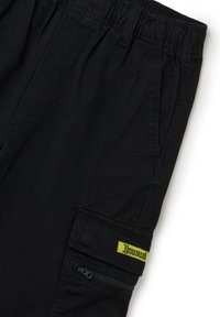 Pantalones cortos de carga negros con cintura elástica. Presentan un bolsillo lateral con cremallera y una etiqueta verde con la palabra "Unusual" cosida.