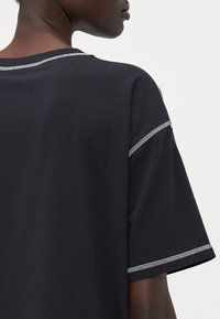 Juodas medvilnės T-shirt su laisvu kirpimu, trumpomis rankovėmis ir kontrastine balta siuvimo linija apykaklėje ir rankovėse.
