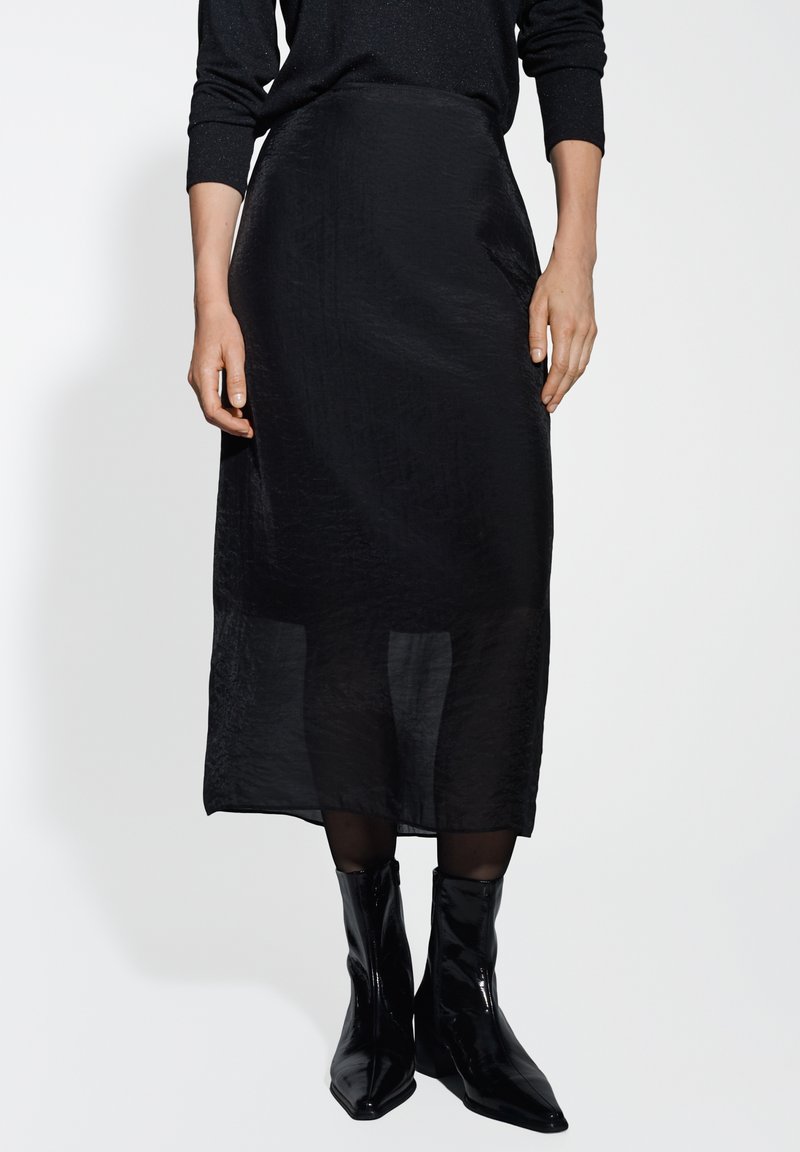 Gonna nera di media lunghezza realizzata in un materiale trasparente e testurizzato. Presenta una silhouette elegante ed è abbinata a stivaletti neri lucidi.