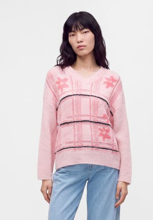 Jeune femme aux longs cheveux noirs porte un pull rose à motifs et un jean bleu clair sur un fond blanc uni.