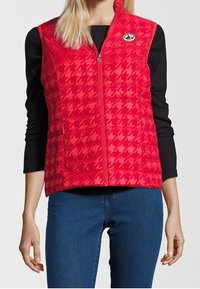 Czerwony pikowany kamizelka z wzorem w piedzie, z zapięciem na zamek błyskawiczny z przodu i logo po lewej stronie klatki piersiowej. Nosi się na czarnej koszuli z długim rękawem.