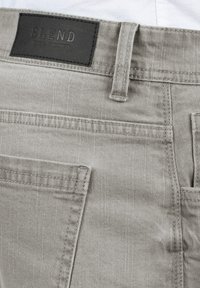 Grå bukser i denimstoff med en teksturert overflate har et lærmerke med teksten "BLEND" over baklommen og synlige sømdetaljer.