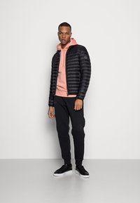 Giacca a vento nera con design trapuntato, indossata sopra un hoodie rosa chiaro, abbinata a pantaloni felpati neri e sneakers nere. Look semplice e moderno.