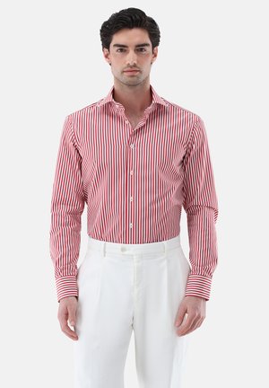 Mann trägt ein rot-weiß vertikal gestreiftes Hemd, das in hoch taillierte weiße Hose gesteckt ist, steht vor einem schlichten Hintergrund.
