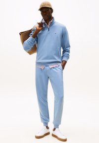 Sweatshirt zippé bleu clair avec col côtelé, associé à un jogging assorti. Des baskets blanches et une casquette beige complètent la tenue.