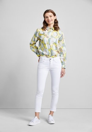 Junge Frau steht lächelnd da, trägt ein bunt gemustertes Langarmshirt, weiße enge Hosen und weiße Sneaker vor einem schlichten Hintergrund.