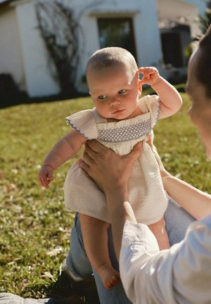 Adult ținând în brațe un bebeluș îmbrăcat într-o rochie albă în aer liber pe iarbă, lângă o clădire albă, într-o zi însorită.