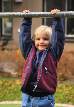 Child hanging from a metal bar, wearing a navy blue and burgundy fleece zip-up jacket with front pockets, and denim jeans.