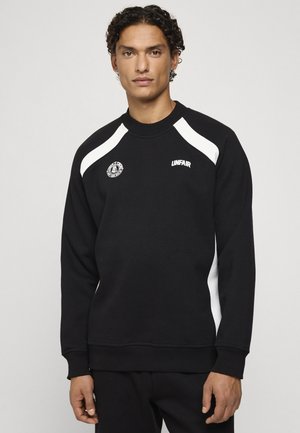Homme aux cheveux bouclés portant un sweat-shirt noir avec des accents blancs sur les épaules et des logos sur la poitrine, debout devant un fond clair uni.