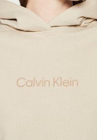 Felpa con cappuccio beige, caratterizzata da una texture a coste, con il logo "Calvin Klein" di un beige chiaro stampato orizzontalmente sul petto in un font sans-serif liscio.