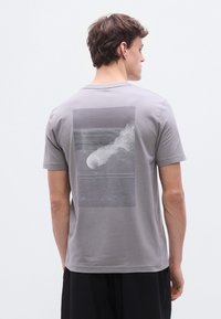 Ανοιχτό γκρι βαμβακερό μπλουζάκι με εκτυπωμένο γραφικό στο πίσω μέρος, που απεικονίζει γραμμές και ένα κυκλικό στοιχείο, κοντά μανίκια και στρογγυλή λαιμόκοψη.