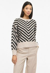 Pull rayé noir et beige avec un motif chevron, texture douce, encolure ronde et poignets côtelés, associé à un pantalon beige clair.