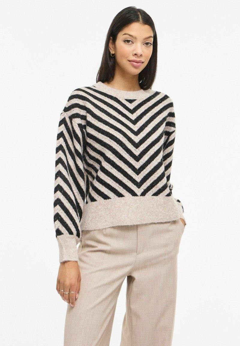 Pull rayé noir et beige avec un motif chevron, texture douce, encolure ronde et poignets côtelés, associé à un pantalon beige clair.