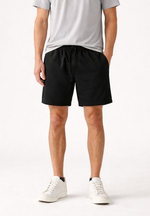 Uomo che indossa pantaloncini neri casual con coulisse, t-shirt grigio chiaro e sneakers bianche, in piedi con le mani nelle tasche su uno sfondo neutro.