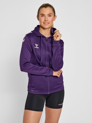Vrouw met blond haar in een paardenstaart, die een paarse hoodie met rits en zwarte sportshorts draagt, staand tegen een effen lichte achtergrond.
