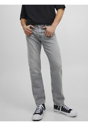 Mann trägt hellgraue Jeans, schwarze Sneakers mit weißen Schnürsenkeln und ein schwarzes Shirt, steht mit Händen in der Nähe der Taschen vor einem grauen Hintergrund.