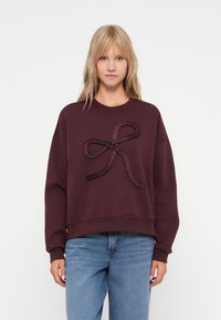 Sweatshirt bordô com um design de fita texturizada na parte da frente, decote redondo, ombros caídos e punhos canelados. Combinado com jeans azuis.