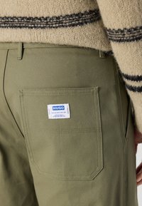 Green cotton trousers with a back pocket featuring a white HUGO label. The fabric has a smooth texture and sturdy construction.