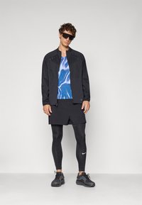Sort letvægtsjakke, blå mønstret tanktop, sorte shorts og sorte leggings. Sorte sneakers, solbriller. Minimalistisk atletisk design.