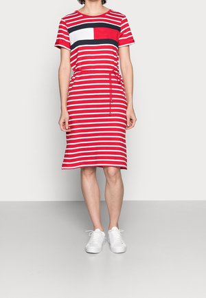 Frau trägt ein knielanges, rot-weiß horizontal gestreiftes Kleid mit einem color-block Brustmuster und weißen Sneakers, steht auf einem grauen Boden.