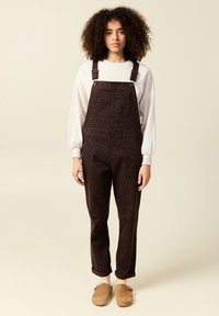 Bruna mönstrade hängselbyxor med justerbara axelremmar, en framficka och uppvikta ärmslut, i kombination med en ljus tröja och beige träskor.