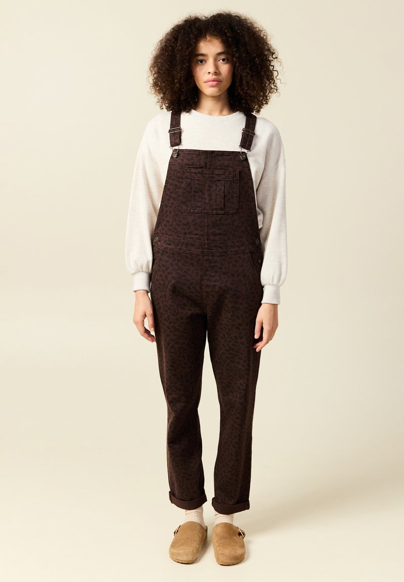 Bruna mönstrade hängselbyxor med justerbara axelremmar, en framficka och uppvikta ärmslut, i kombination med en ljus tröja och beige träskor.