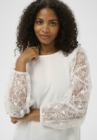 Haut blanc avec manches transparentes brodées de motifs floraux. Les manches sont bouffantes et présentent des motifs floraux blancs délicats. Encolure ronde simple.
