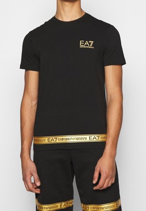 Man draagt een zwart Emporio Armani EA7 T-shirt en bijpassende korte broek met gouden logo banden aan zoom en borst.