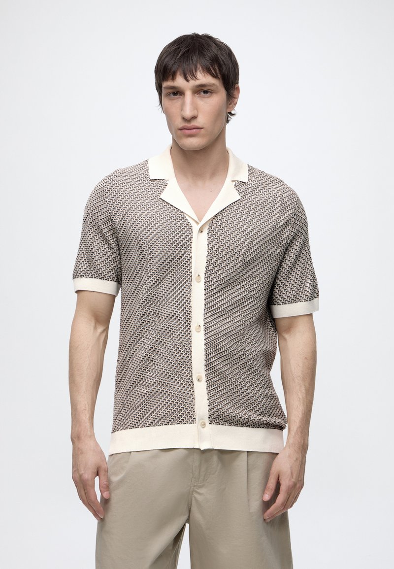 Jeune homme portant une chemise à manches courtes, boutonnée, à motifs avec un bord clair et un pantalon beige, debout devant un fond uni.
