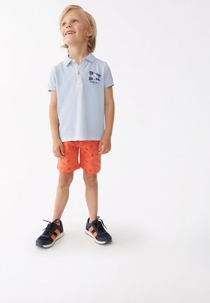 Camisa polo azul clara con un logo, pantalones cortos naranjas con pequeños dibujos y zapatillas azul marino con acentos naranjas. Niño de pie, sonriendo.