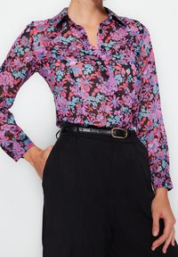 Blusa de manga larga con botones en tejido negro translúcido, con un patrón floral en rosa, púrpura y azul. Combinada con pantalones negros de cintura alta.
