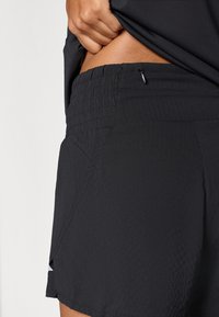 Pantalones cortos negros con una tela texturizada, cintura elástica, bolsillos laterales y un pequeño detalle de cremallera en la parte trasera para un almacenamiento seguro.