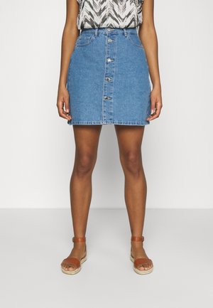 Spódnica dżinsowa z guziczkami z przodu, jasne pranie, kieszenie na biodrach i postrzępiony brzeg. Połączona z brązowymi sandałami na paski. Stylizowana z wzorzystą bluzką.