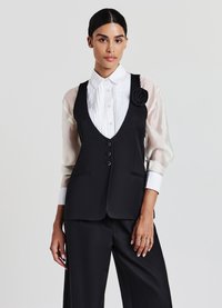 Schwarzes tailliertes Weste mit tiefem V-Ausschnitt, ausgestattet mit drei Knöpfen und Seitentaschen. Kombiniert mit einem weißen plissierten Hemd und transparenten Ärmeln.