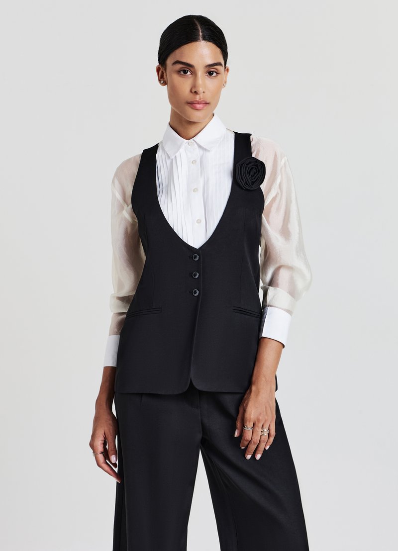 Schwarzes tailliertes Weste mit tiefem V-Ausschnitt, ausgestattet mit drei Knöpfen und Seitentaschen. Kombiniert mit einem weißen plissierten Hemd und transparenten Ärmeln.