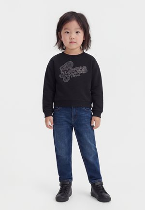 Bambino con capelli neri fino alle spalle che indossa una felpa nera con il logo "Guess", jeans blu e sneakers nere alte, in piedi rivolto in avanti.
