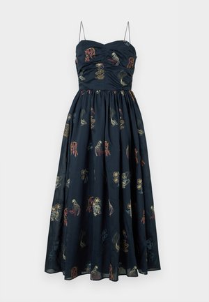 Femme aux cheveux foncés portant une robe midi bleu marine à fines bretelles et motifs abstraits multicolores, associée à des talons pointus blancs.