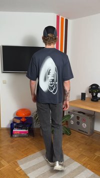 T-shirt oversized cinzento escuro com uma estampa gráfica abstrata branca nas costas. Modelo a usar calças cinzentas e ténis, num ambiente interior.